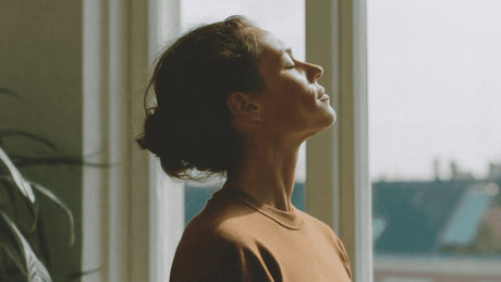 Woman standing by a window with eyes closed, enjoying soft morning sunlight on her face