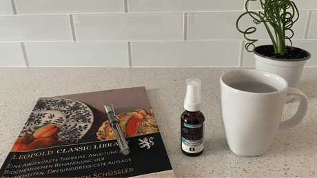 AnnaKare Mag Phos cell salt spray on a kitchen counter beside warm water, a pen, and an old German Schuessler cell salts book.