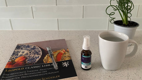 AnnaKare Mag Phos cell salt spray on a kitchen counter beside warm water, a pen, and an old German Schuessler cell salts book.