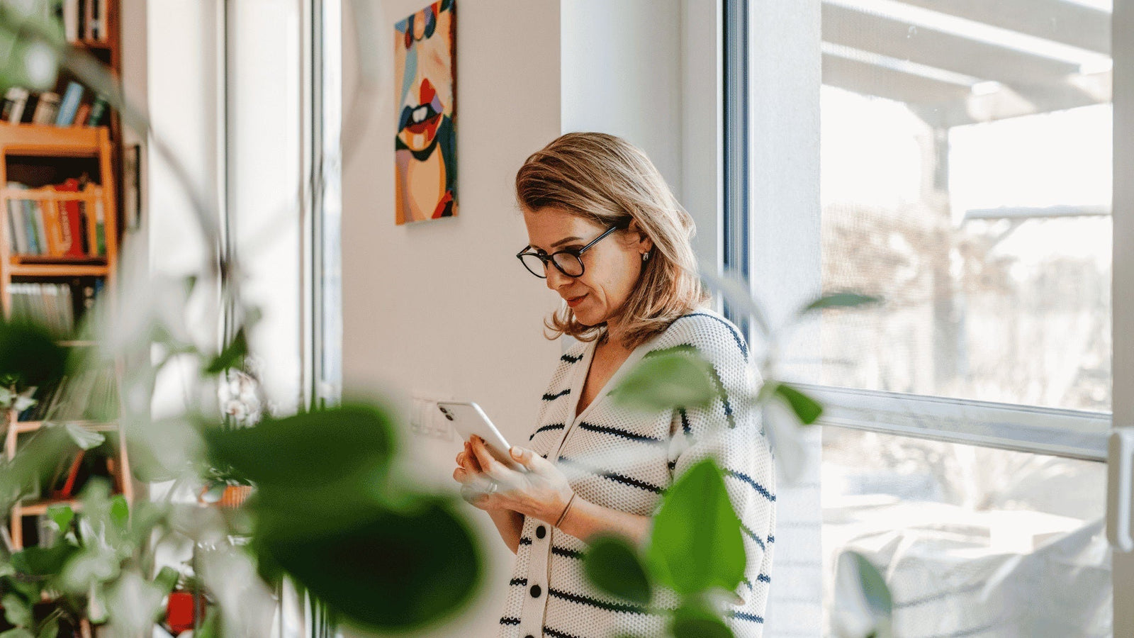Middle-aged woman leaning against a living room window, looking at information on her phone about perimenopause and menopause wellness.