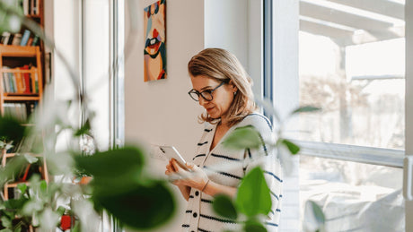 Middle-aged woman leaning against a living room window, looking at information on her phone about perimenopause and menopause wellness.